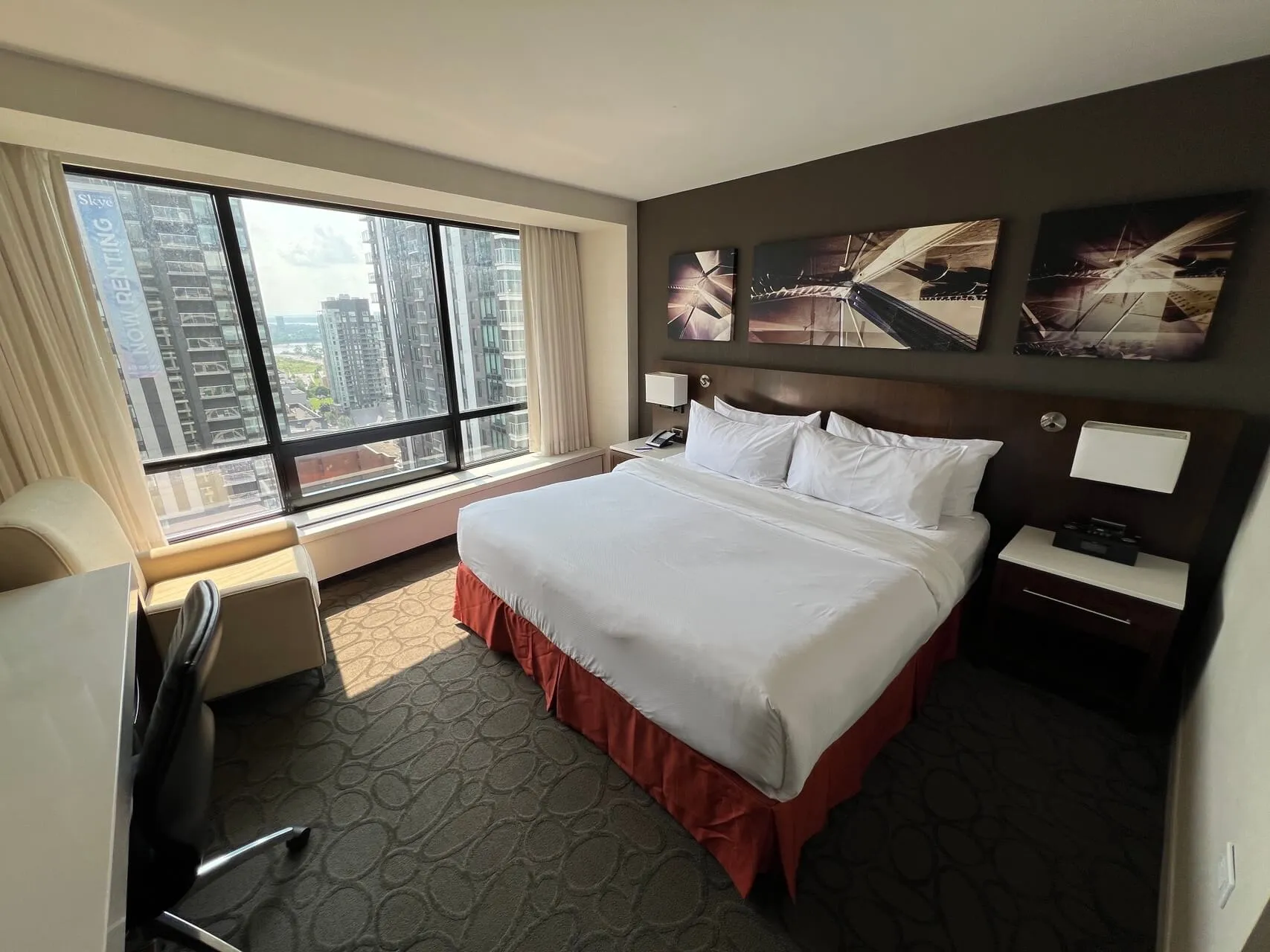 Queen bed with red skirt and city skyline view from high-rise hotel room with floor-to-ceiling windows